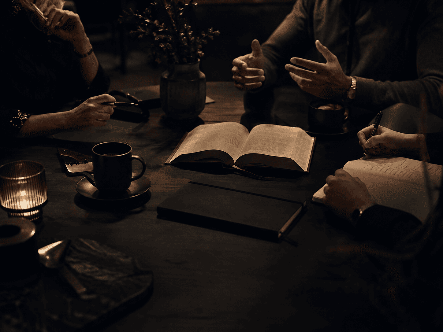 Pessoas estudando ao redor de uma mesa com um livro aberto, cadernos e café